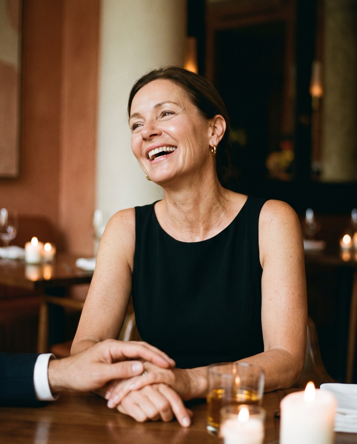 A woman in her fifties laughing at dinner, a hand resting on hers